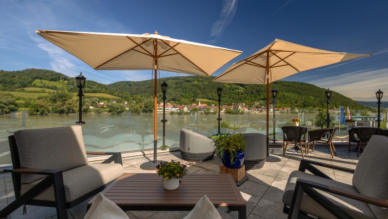 Terrace with a view of the Danube and surrounding hills, equipped with parasols and seating furniture.