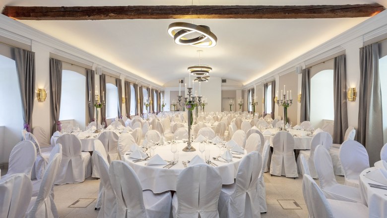 A festively decorated hall with round tables, white tablecloths and chair covers, candlesticks and modern lighting.