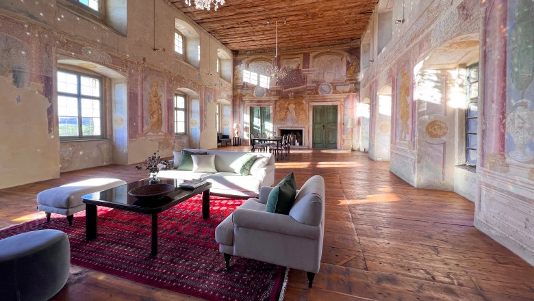 A historic ballroom with a wooden floor, painted walls and a chandelier. Modern furniture and a red carpet are placed in the room.