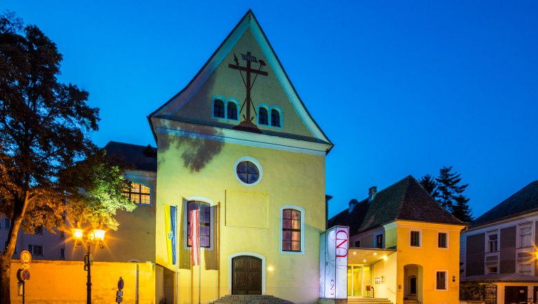 Yellow building with cross symbol, illuminated at night.