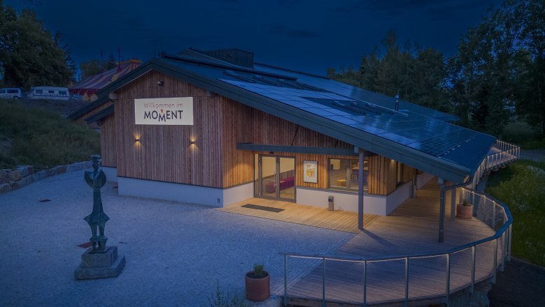 A modern building with wooden cladding and solar panels on the roof, illuminated at dusk. A sign with the inscription 'Welcome to MOMENT' is visible.