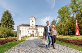 A couple walks along a cobbled path in front of the Schlosshotel Rosenau, surrounded by green trees and an Austrian flag.