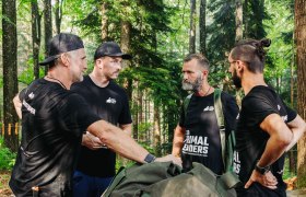 Four men in black T-shirts are standing in the forest and talking.