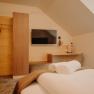 A cozy hotel room photographed from the bed. A flat-screen TV hangs on the wall across from the bed. Next to it is a light-wood wardrobe; part of the wall is also paneled with wood. There is also a shelf and a desk with a chair and a lamp. The room has a sloped ceiling.