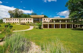 A modern building with large windows and a terrace, surrounded by a well-tended garden and trees under a blue sky.
