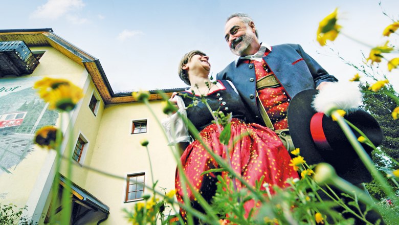 A couple in traditional costume stand in front of a yellow building surrounded by flowers, smiling at each other.