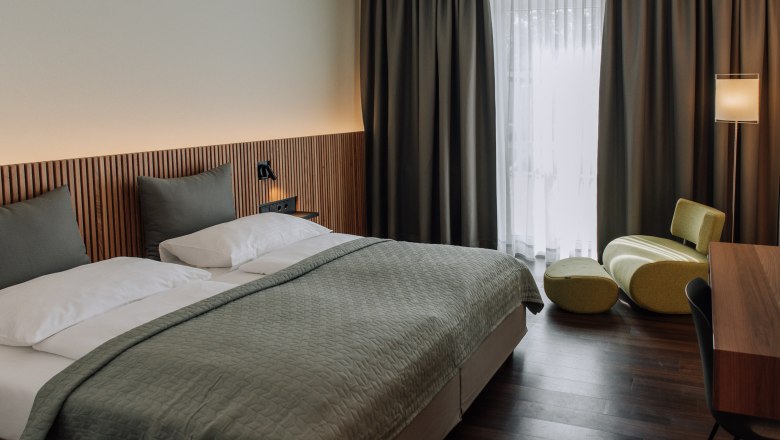 Modernly furnished hotel room with double bed, green armchair and desk.