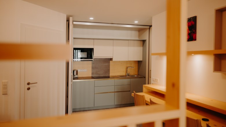 A modern kitchenette in an apartment, decorated in light colors. It features a sink, a microwave, a large stovetop, and an electric kettle. The kitchen can be hidden behind a sliding partition. In front of the kitchen, there is a table against the wall and four armchairs.