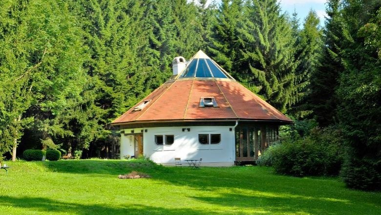A round, white house with a pointed roof in the middle of a green forest.