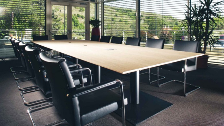 Modern seminar room with large wooden table and black chairs, surrounded by glass walls with a view of the green landscape.