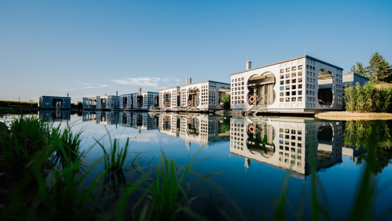 Modern villas on the water with reflections, surrounded by nature.