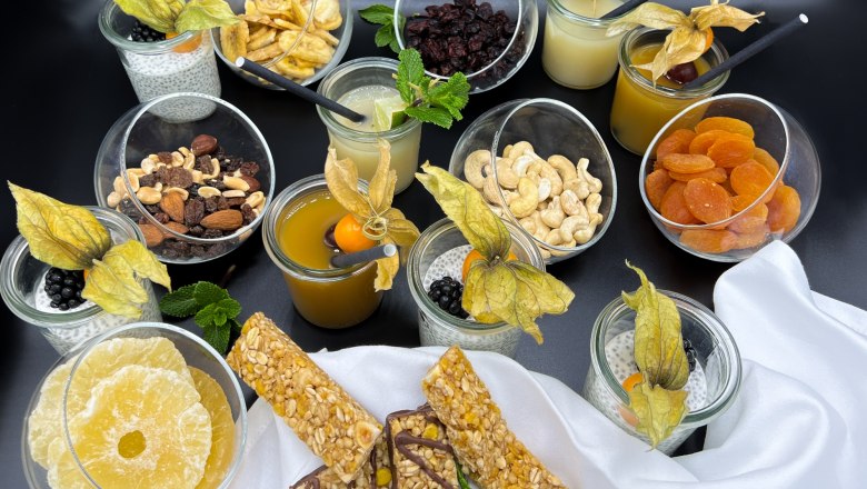 A selection of snacks and drinks, including nuts, dried fruit, chia pudding, and juices, arranged on a table.